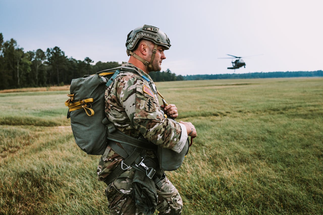 A soldier in camouflage walking in a field with a helicopter overhead.