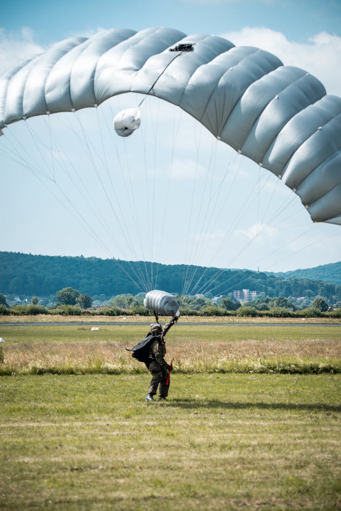 A paratrooper in full gear landing safely on a field under a large parachute during a military exercise.