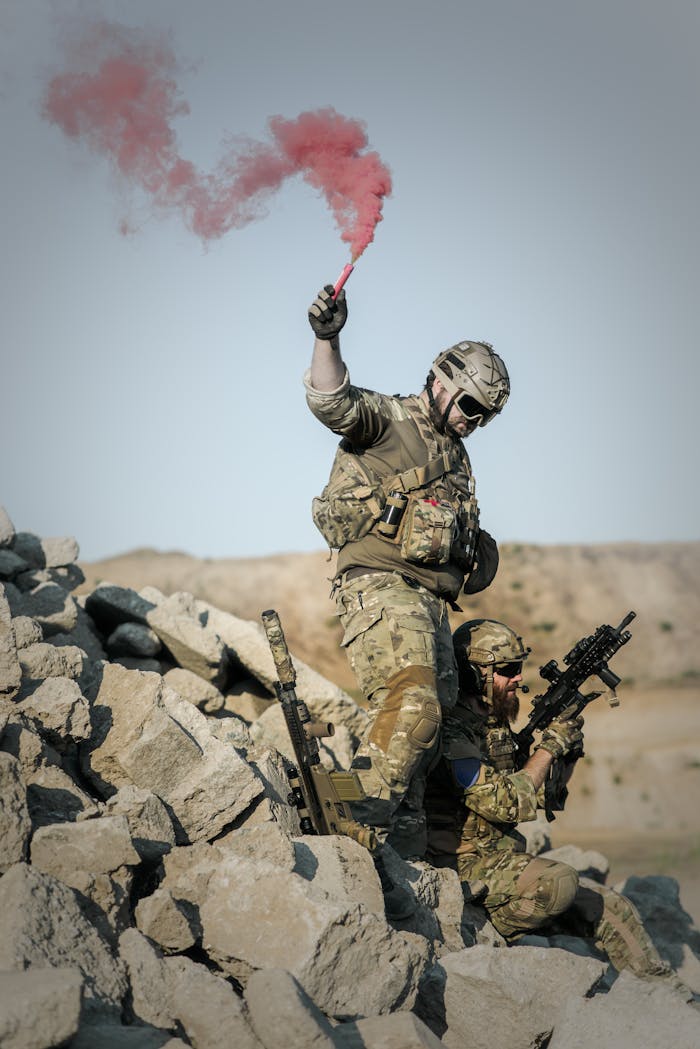 Soldiers in camouflage conduct a military operation using a red smoke signal.